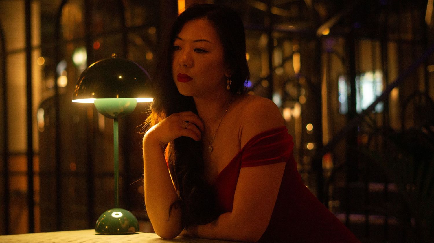 Woman in red off-shoulder dress poses thoughtfully beside a glowing green lamp at a dimly lit bar.