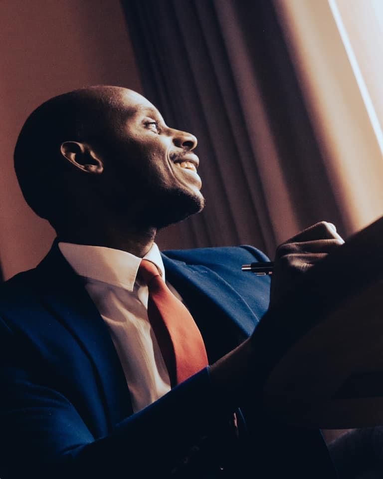 A smiling Black man in a navy blue suit and red tie holds a pen and notepad, looking upward confidently.