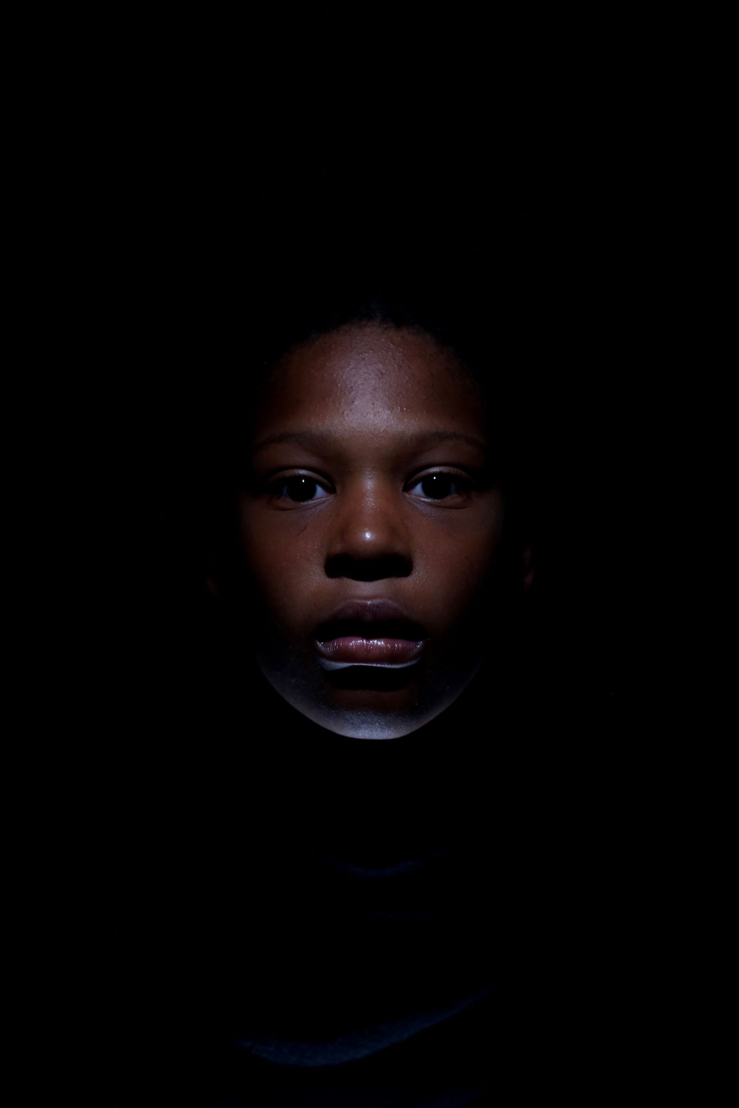 Dramatic low-key portrait of a young person illuminated against a dark black background.