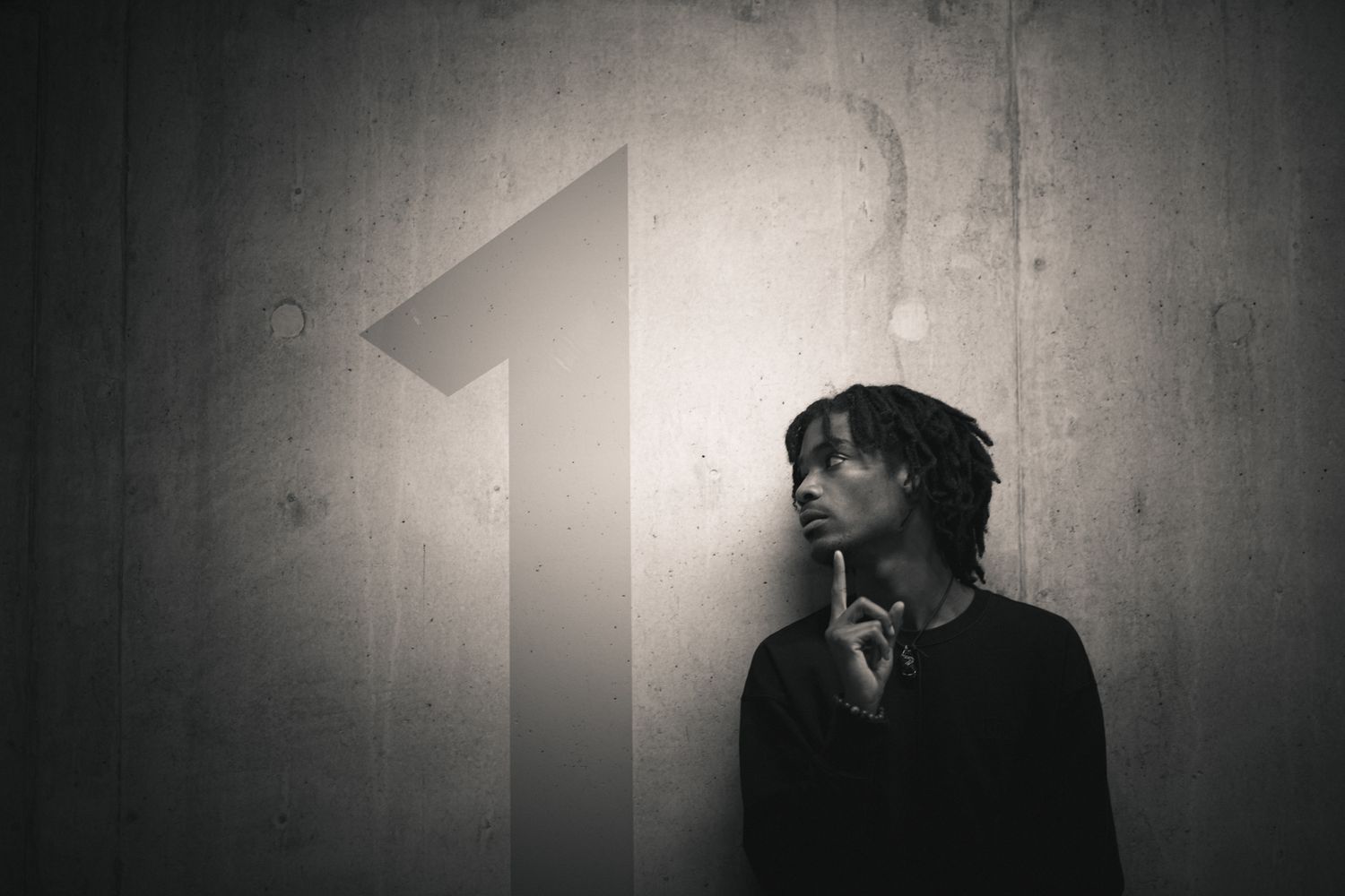 Black and white portrait of a young man with dreadlocks posing thoughtfully beside a large number one on a wall.