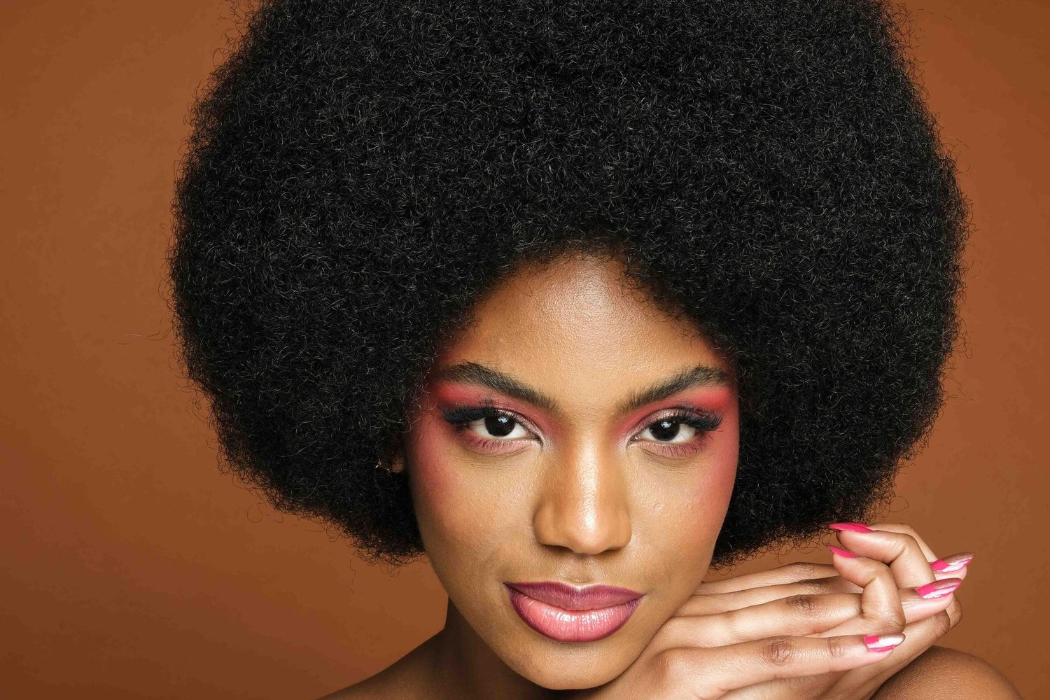 Woman with large natural afro hairstyle wearing bold pink eye makeup and mauve lipstick against a brown background.