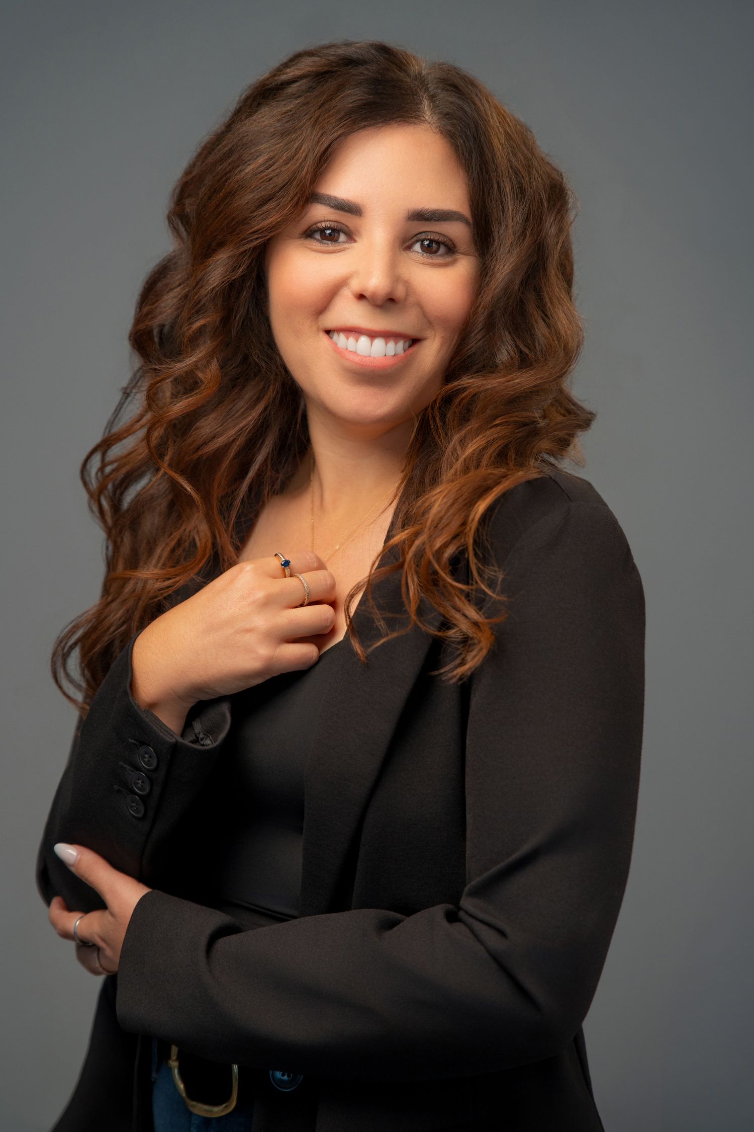 Smiling woman with curly brown hair wearing a black blazer poses confidently against a gray studio background.