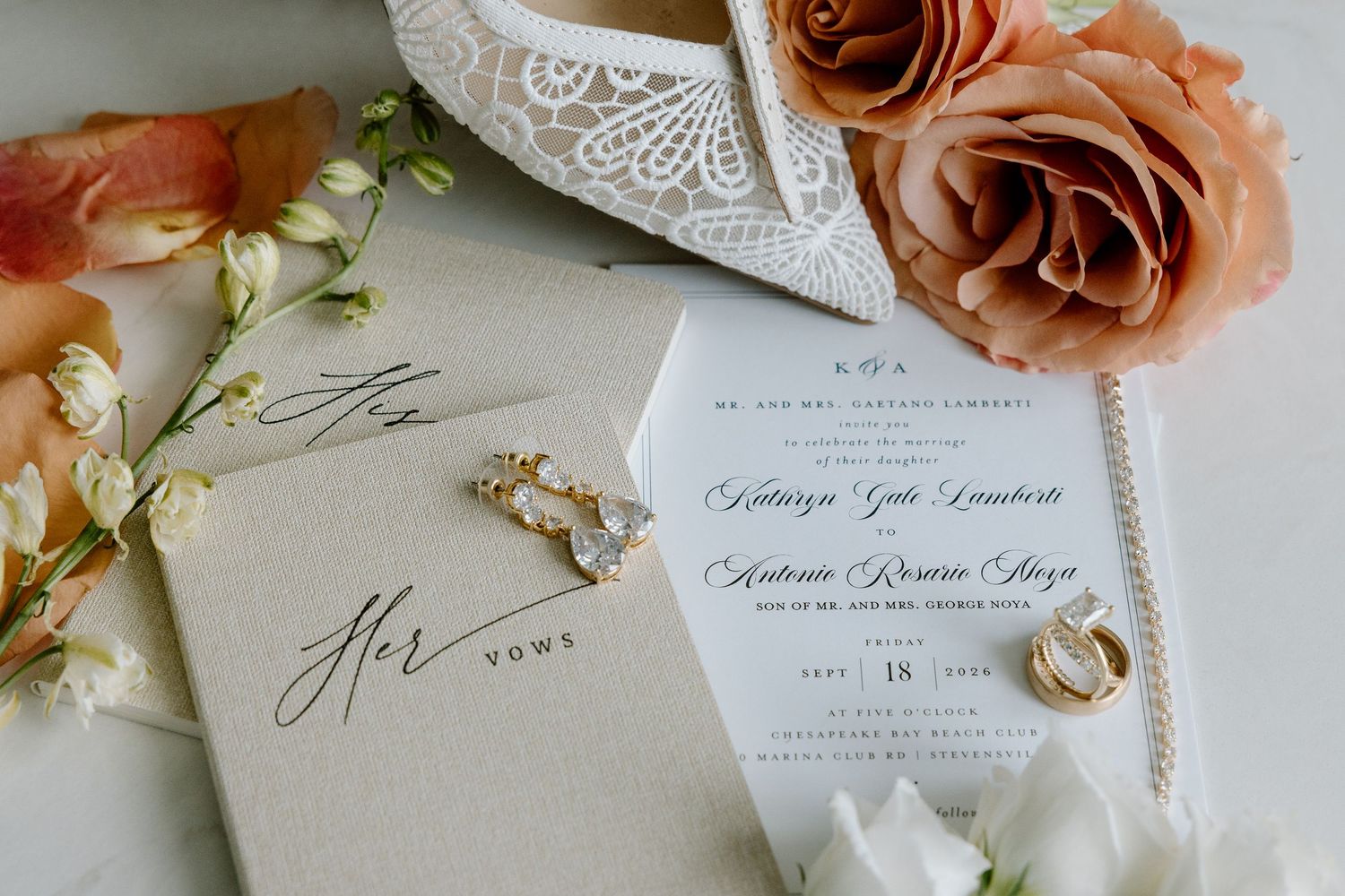 White lace bridal heels adorned with peach roses and white flowers on a marble surface.