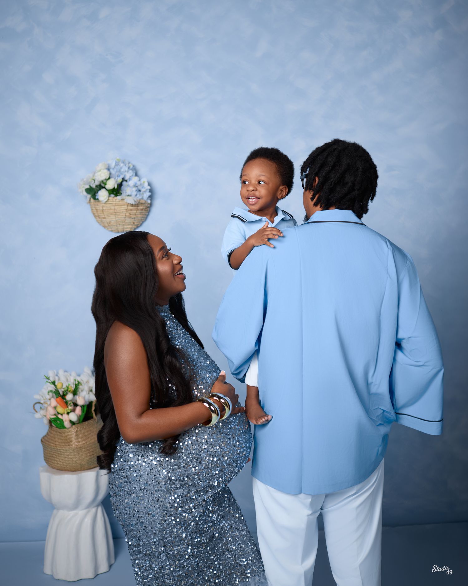 A pregnant Black woman in a blue sequin dress poses with her partner in a light blue suit holding their smiling toddler.