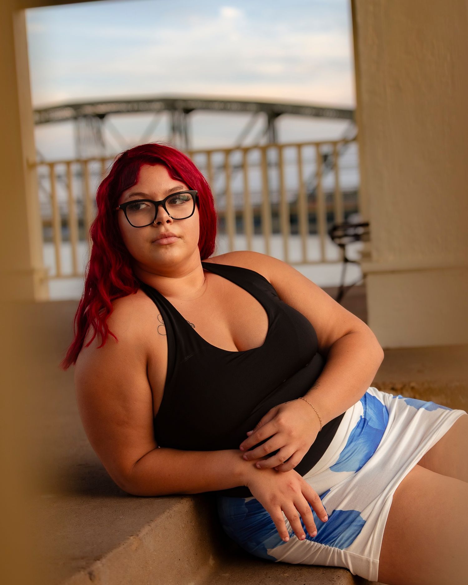 Young woman with red hair and glasses in black top and blue skirt poses near a bridge at golden hour.