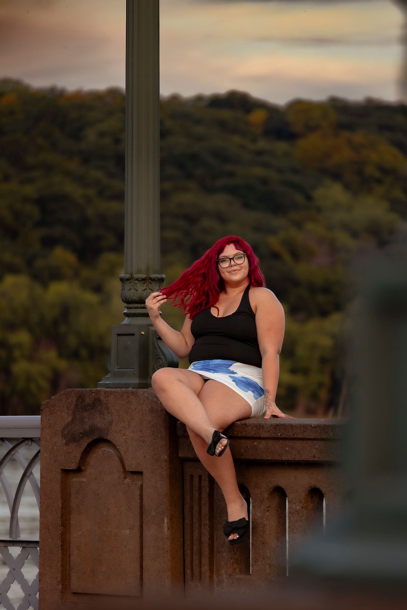 Woman with red hair and glasses sits on stone bridge railing wearing black top and denim skirt at sunset.