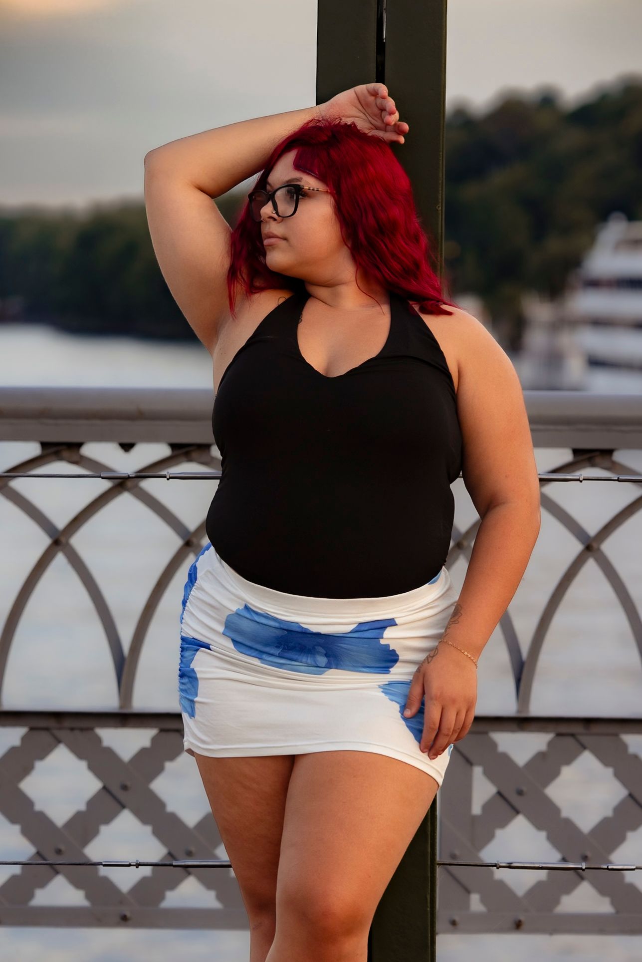 Plus-size woman with red hair and glasses poses on a bridge in a black top and white blue-print mini skirt.