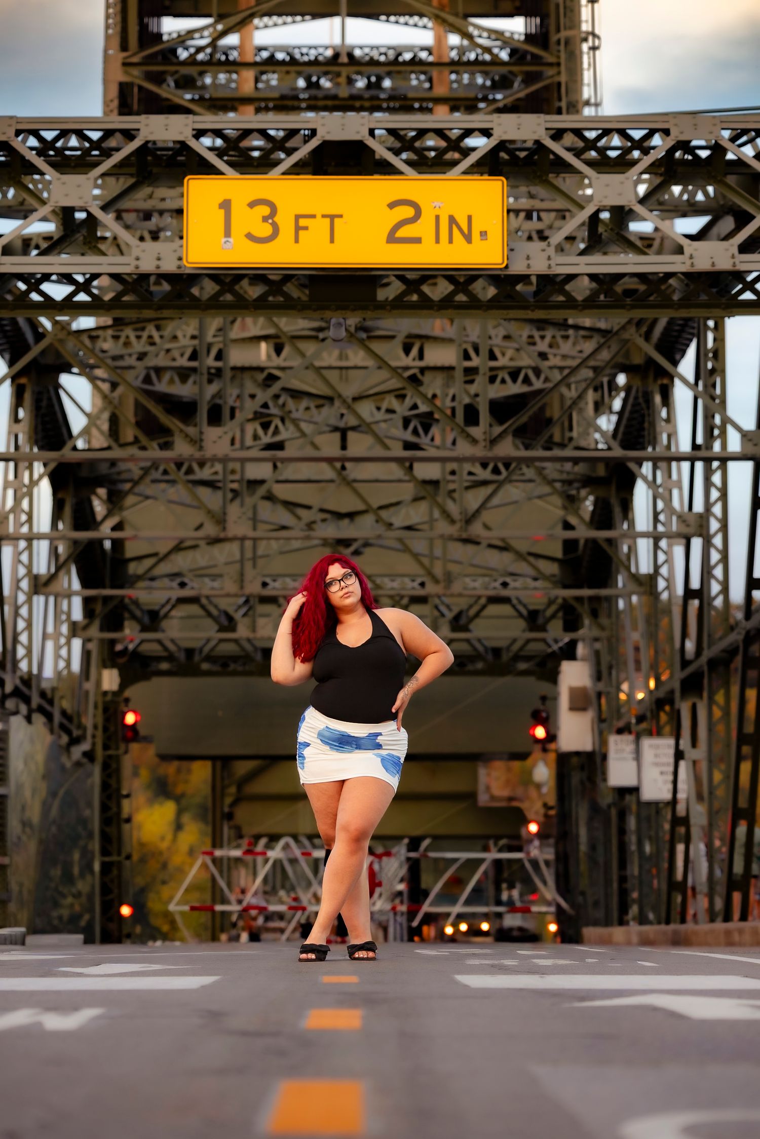 Woman with red hair in black top and denim shorts poses on bridge road under 13ft 2in clearance sign.