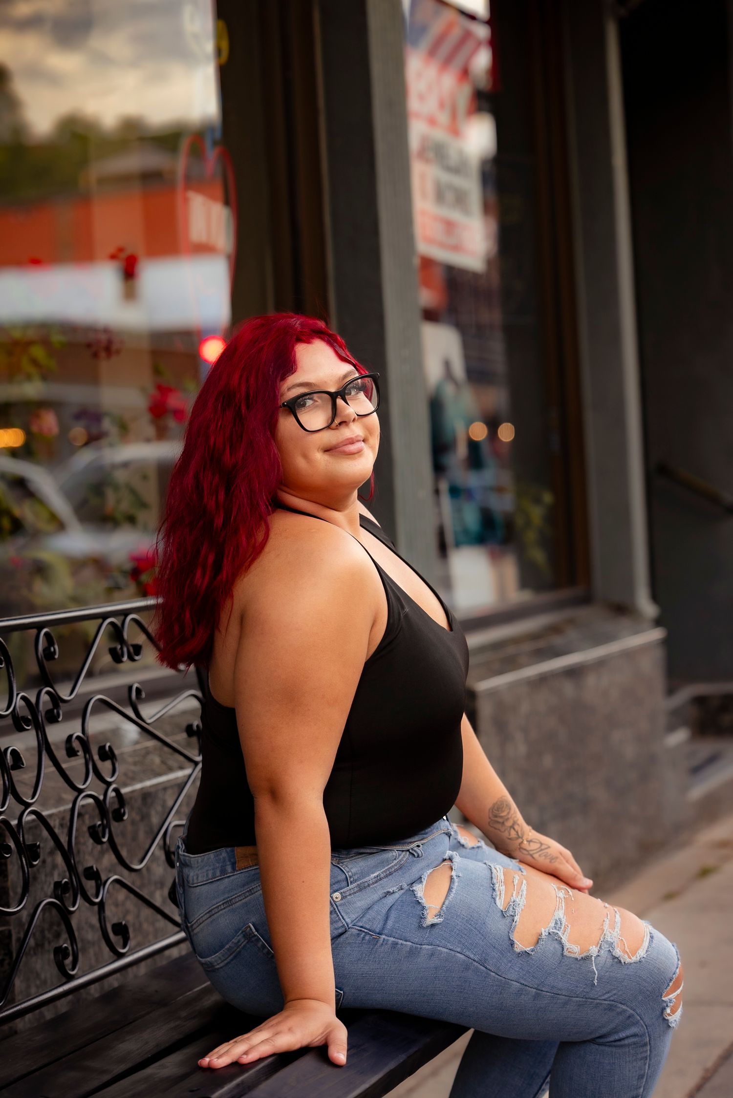 A plus-size woman with red hair and glasses sits on a bench in ripped jeans and black top outside a storefront.