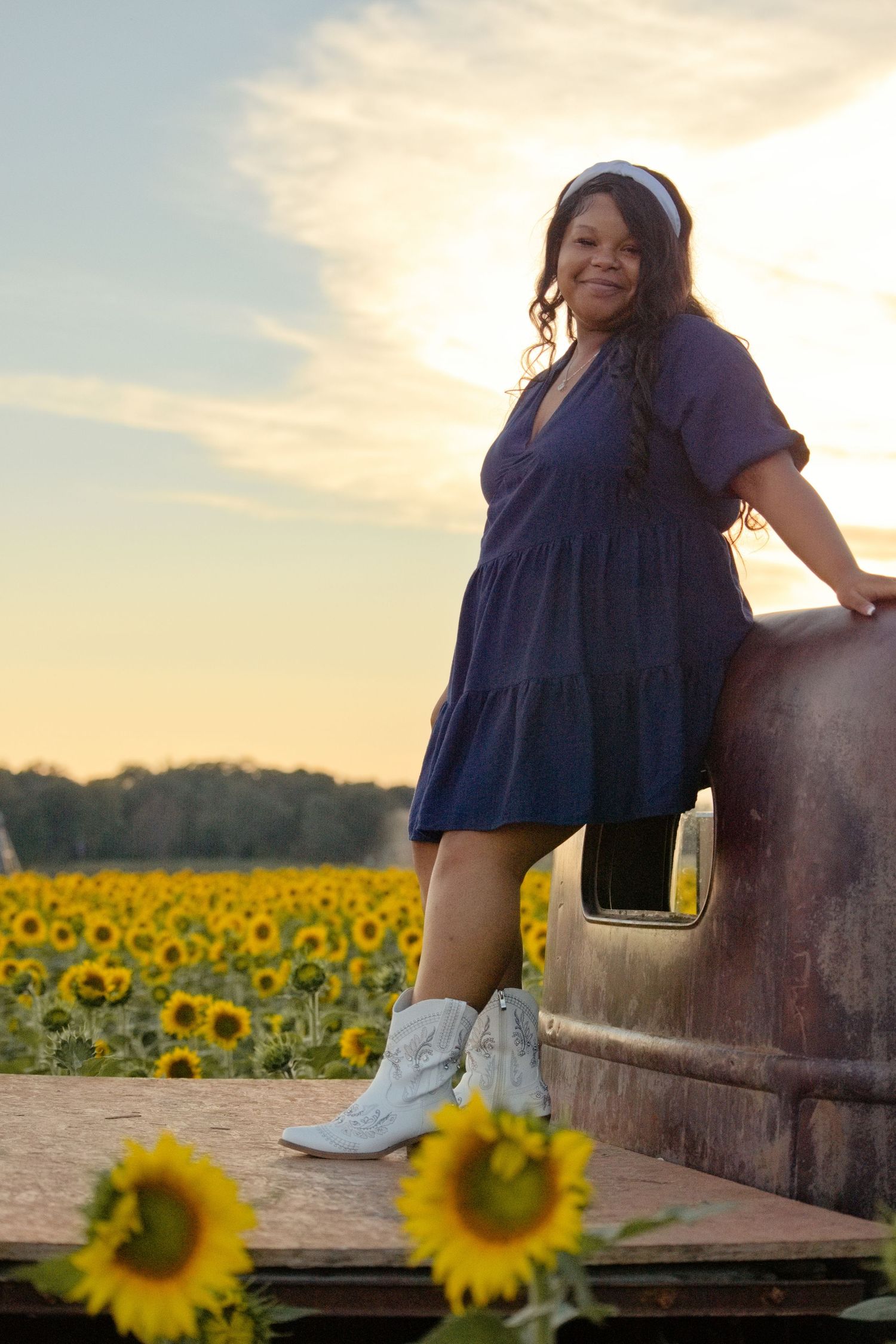 Woman in blue dress and white cowboy boots leaning on vintage truck in sunflower field at sunset.