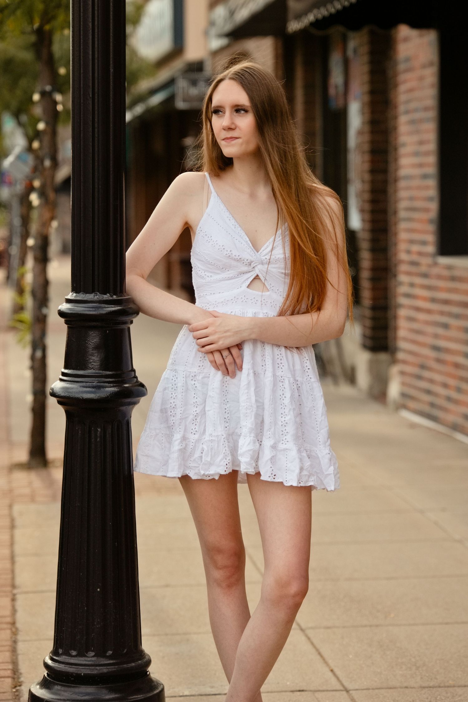 Young woman in white sundress posing by black street lamp on brick sidewalk.
