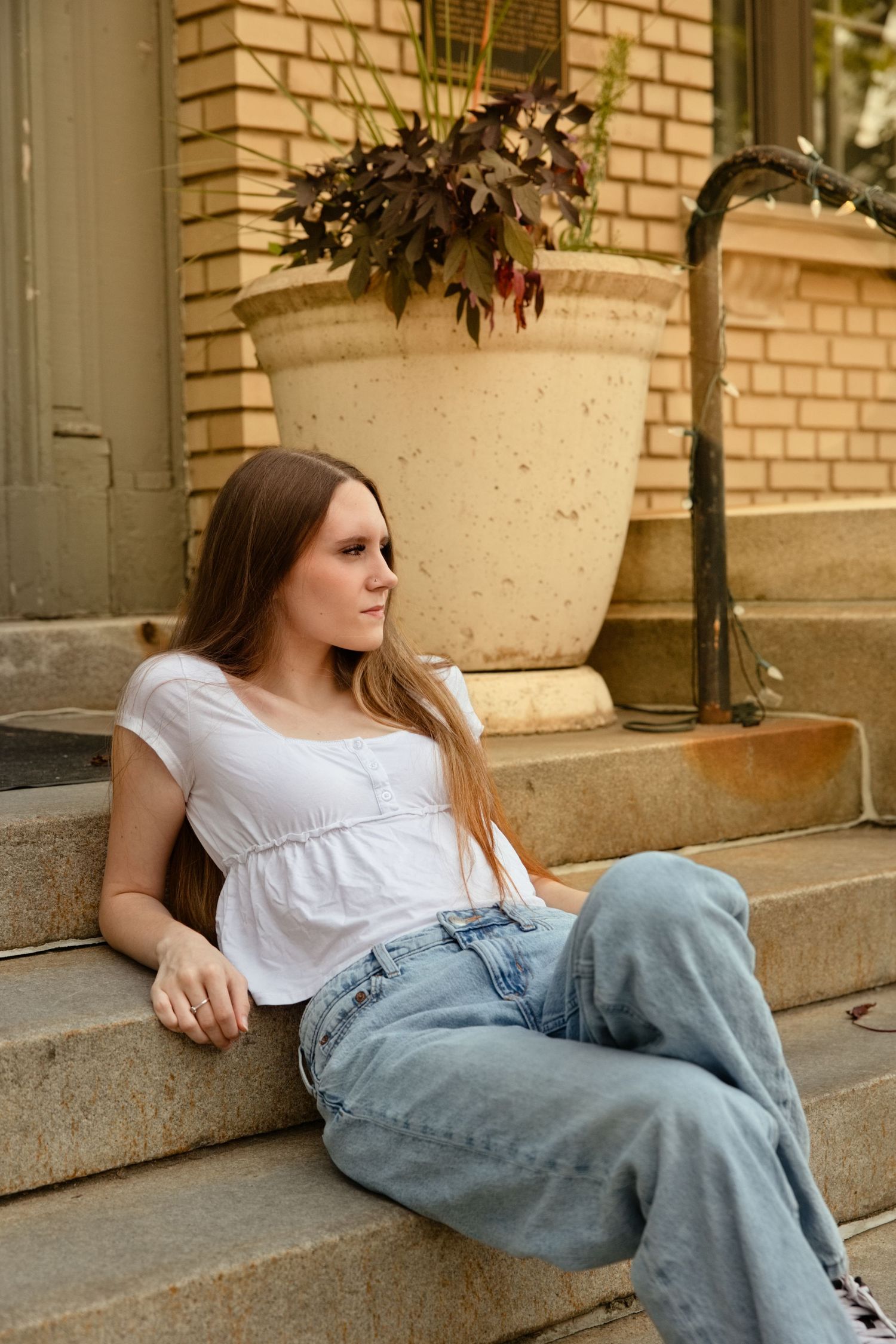 Young woman in white top and jeans sitting relaxed on stone steps outside a brick building with a large planter.