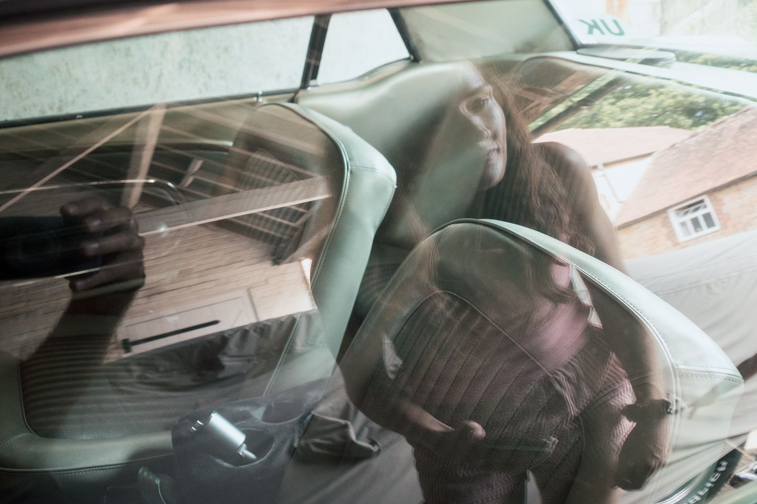 Reflective view through car window showing woman with long hair sitting in driver seat of a vintage vehicle.