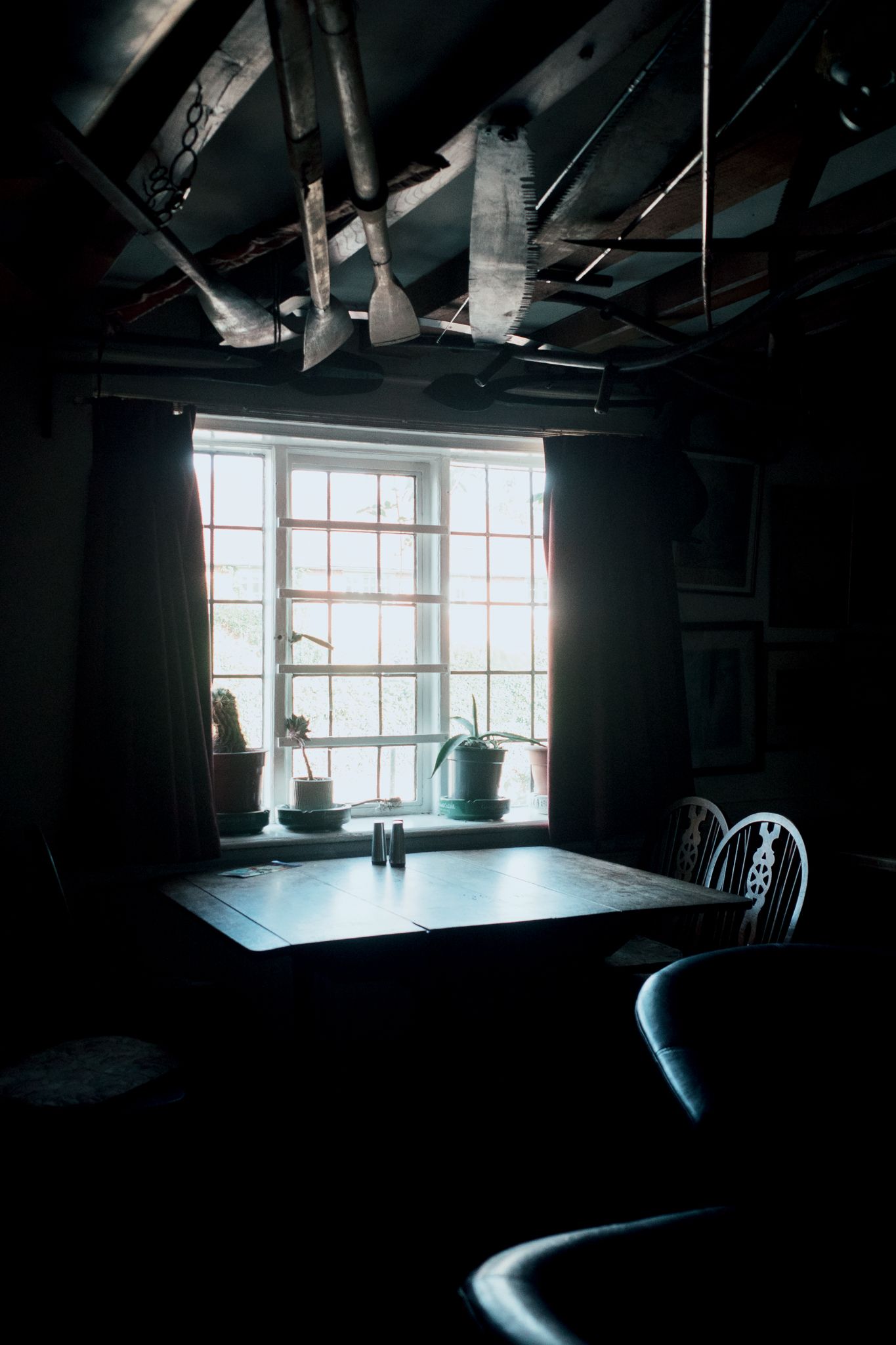 Dark moody interior room with a wooden table, white chairs, potted plants, and bright window casting dramatic light.