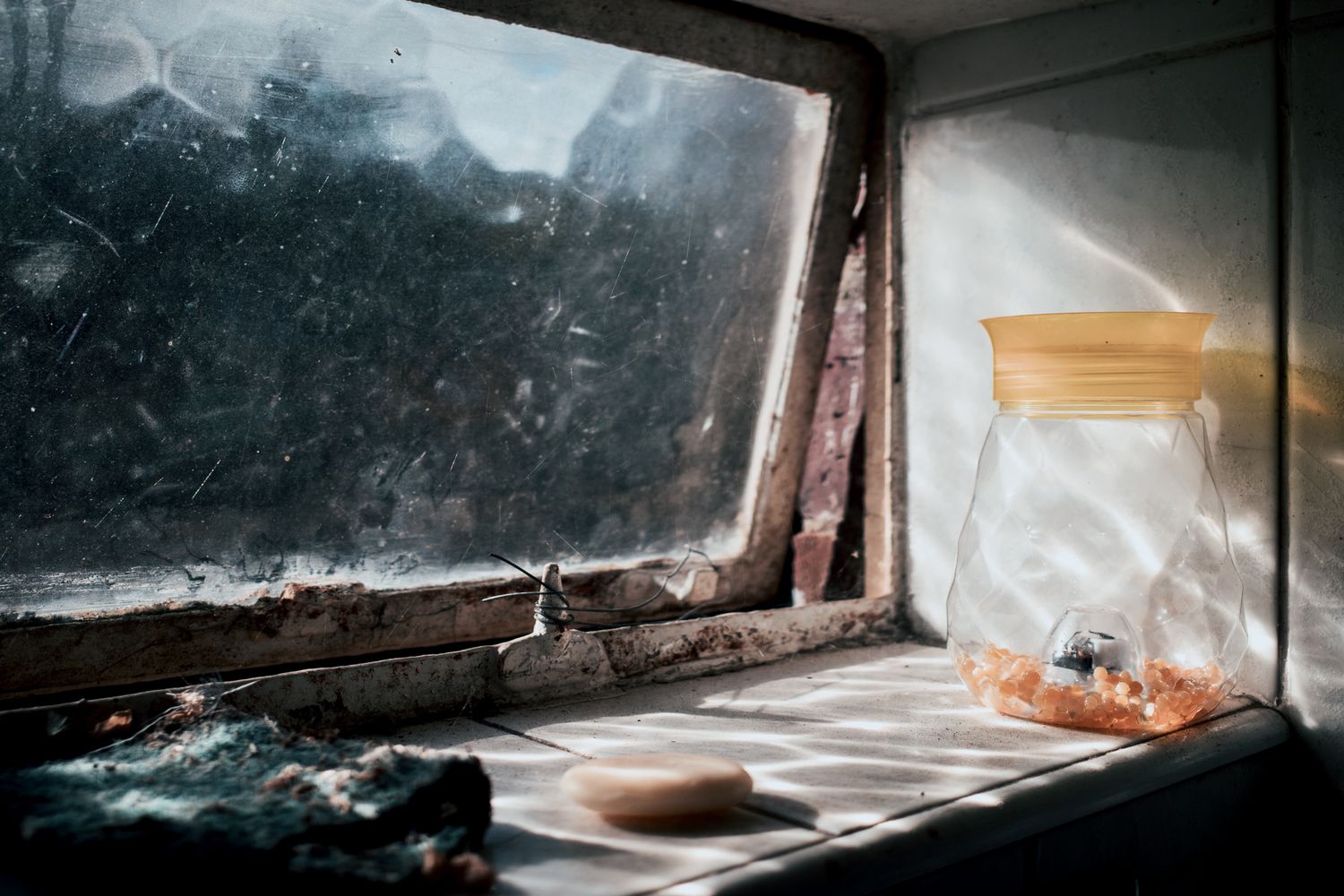 A dusty windowsill with an open glass jar and soap bar, soft light filtering through a grimy, weathered window.