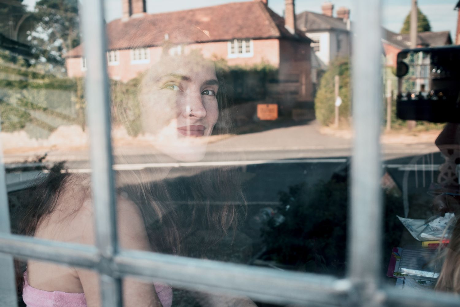 A young woman gazes through a grid window, with a blurred village street reflected in the glass behind her.