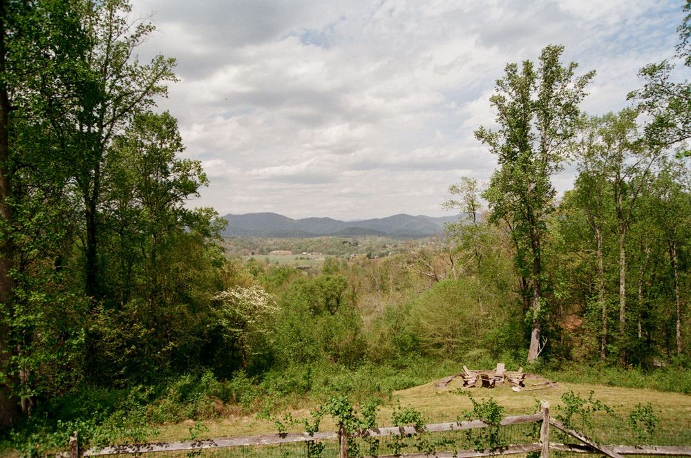 Scenic overlook of a lush green valley with rolling mountains under a cloudy sky, framed by tall trees.