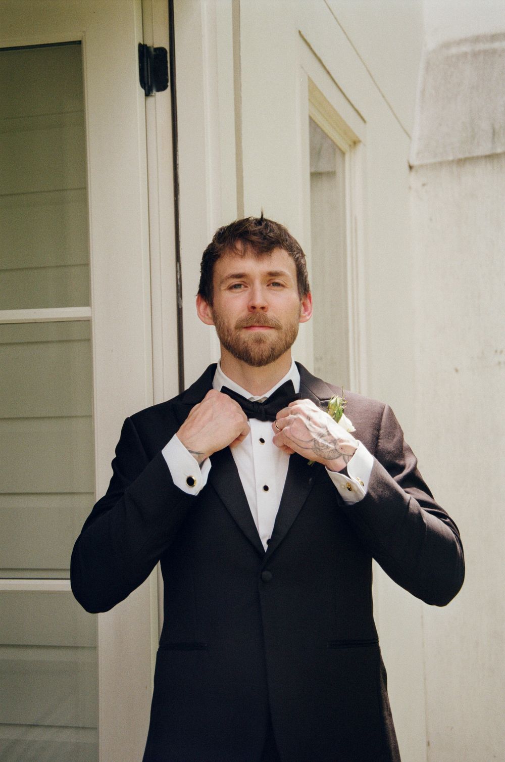 Groom in black tuxedo adjusting bow tie with floral boutonniere, standing outdoors near white building doors.