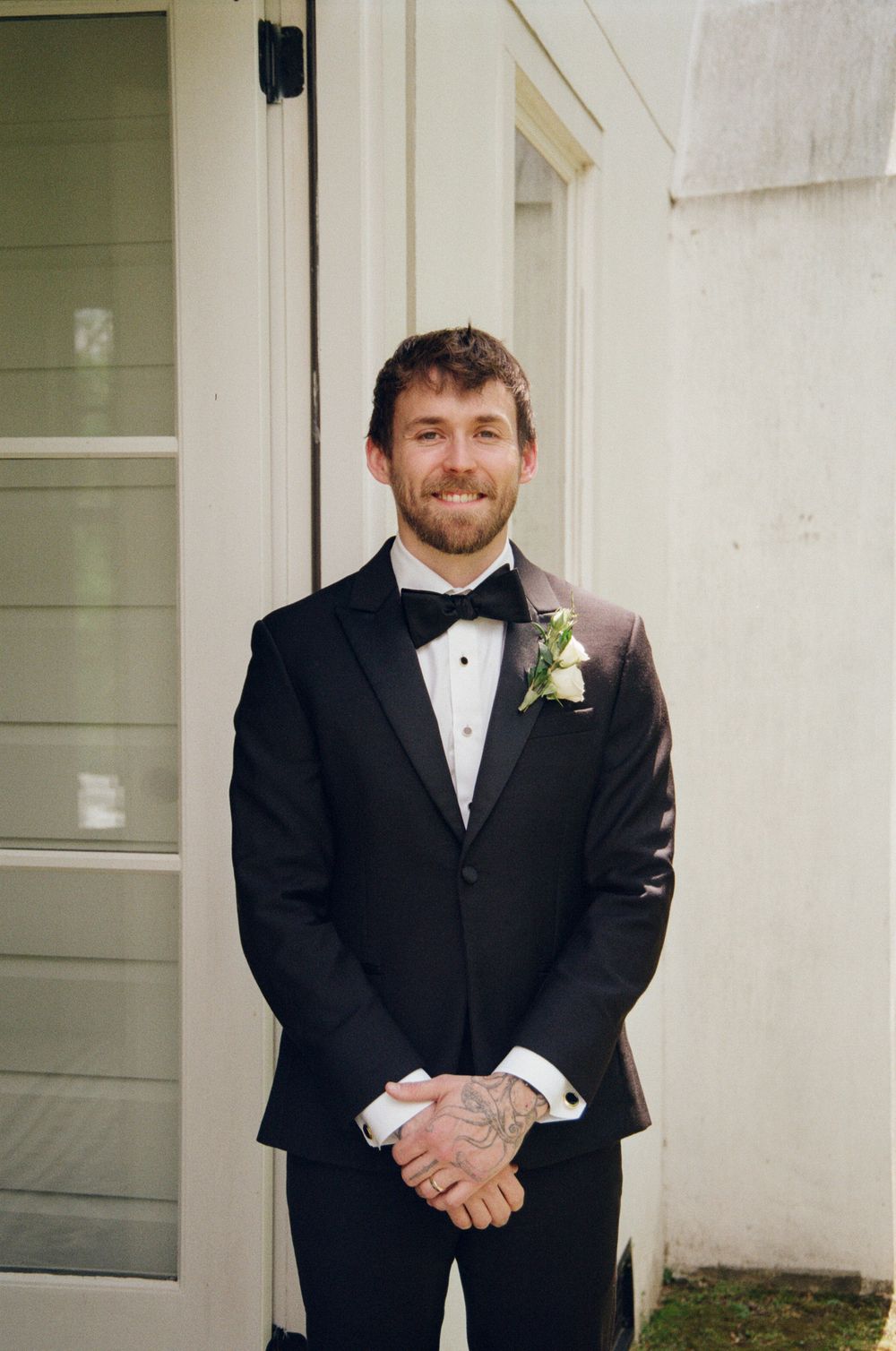 Smiling groom in black tuxedo with bow tie and white boutonniere stands by white door.