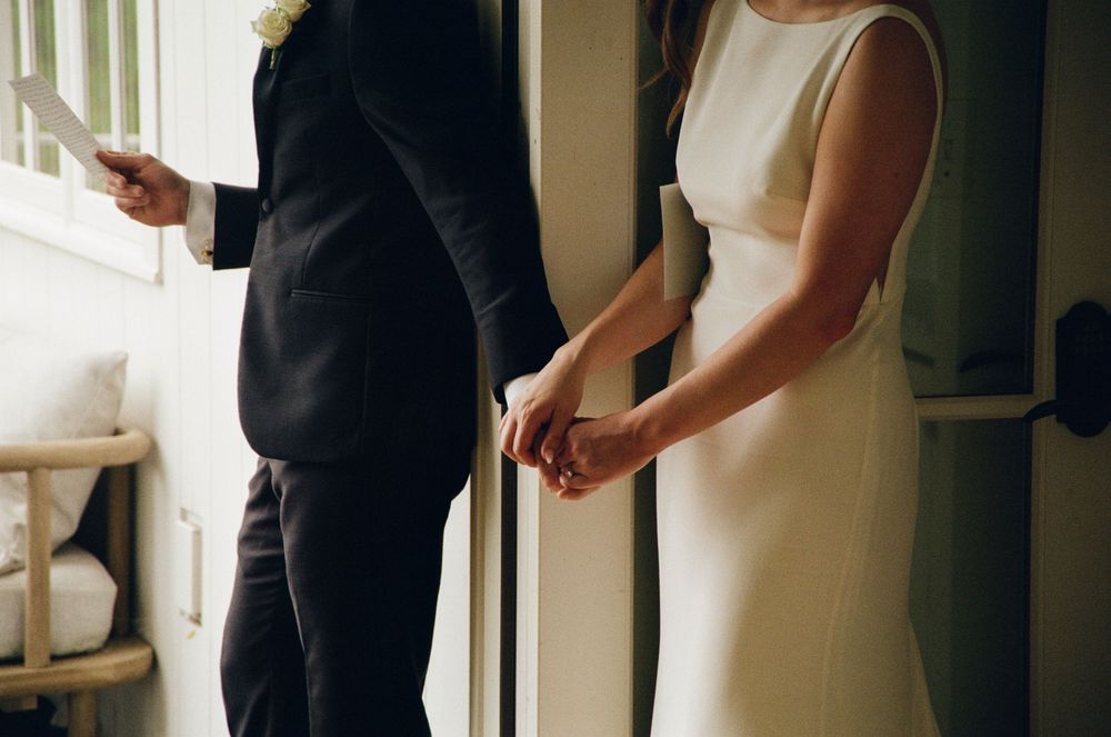 Bride and groom holding hands in elegant wedding attire, sharing a romantic moment by a window.