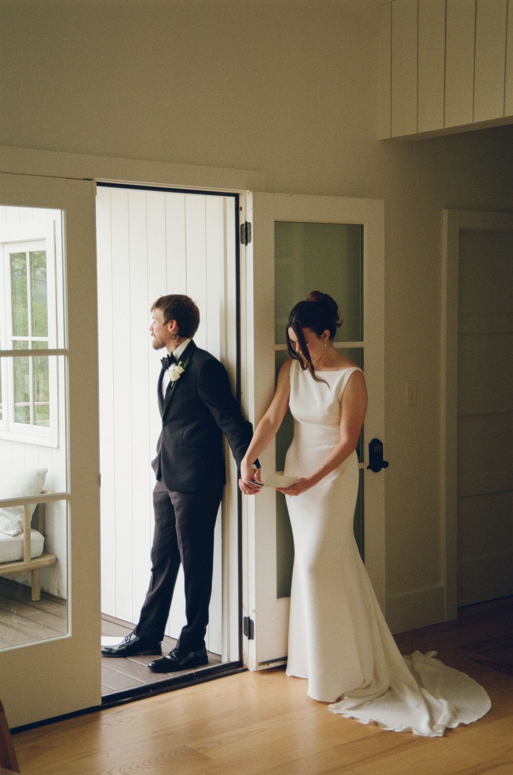 Wedding couple in elegant attire hold hands while standing at open white door in rustic interior.