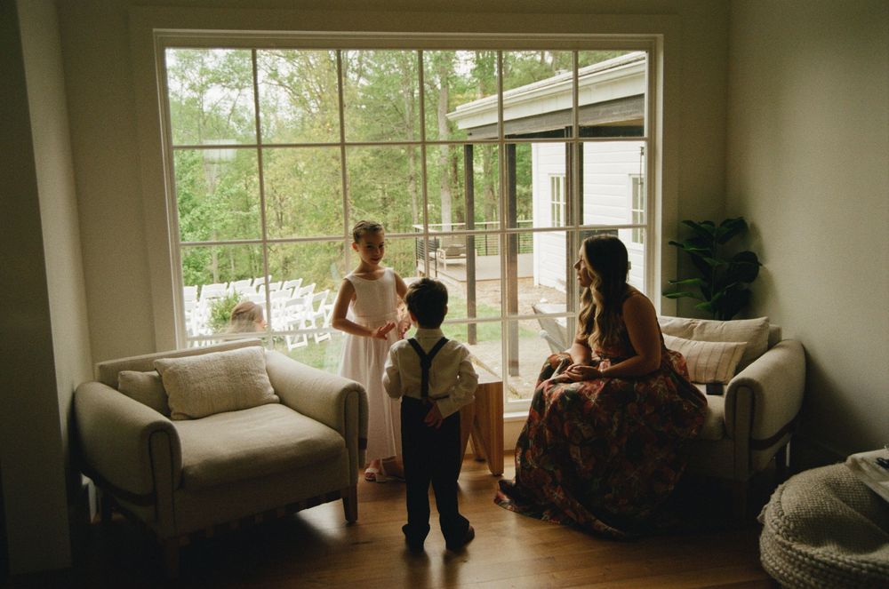 A bride in a floral dress sits with two children by a large window before a wedding ceremony.