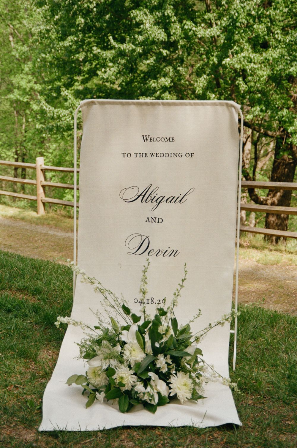 Elegant outdoor wedding welcome sign for Abigail and Devin with white floral arrangement on green lawn.