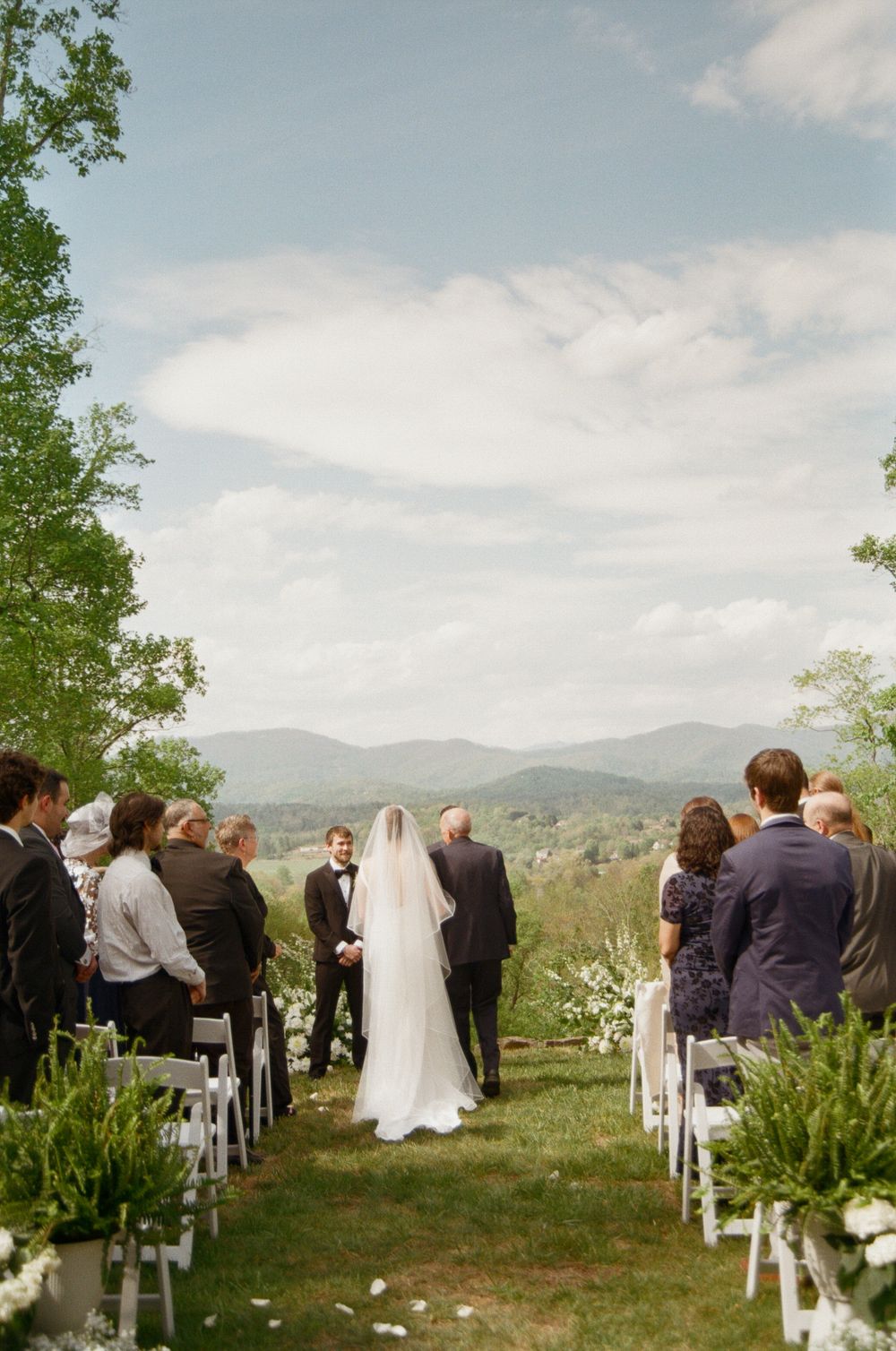 Bride walking down outdoor aisle with mountain views, white chairs, and lush greenery at a scenic wedding ceremony.