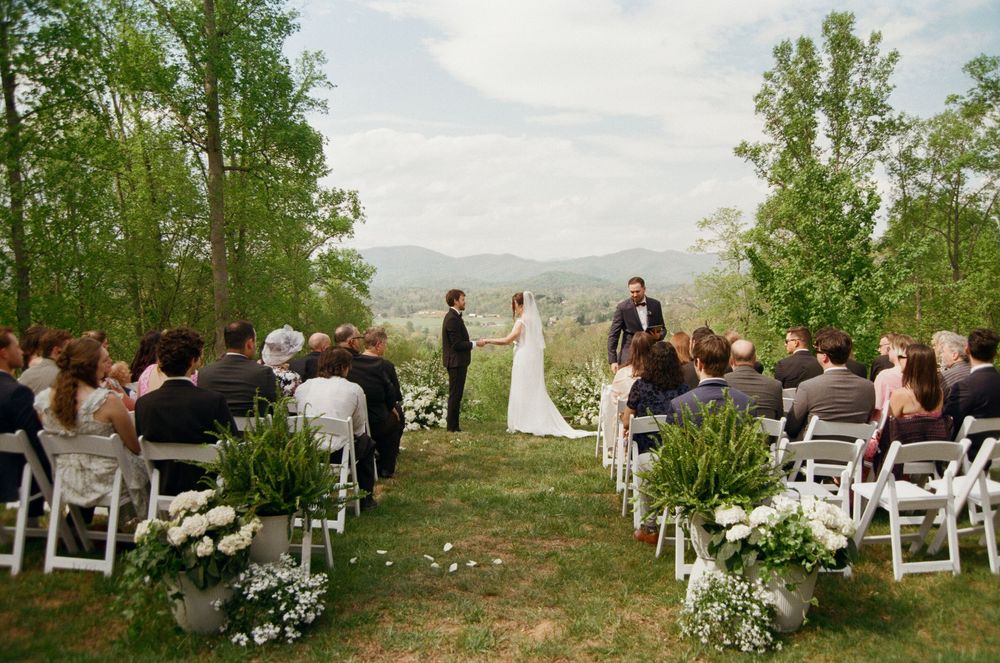 Outdoor wedding ceremony on a green hillside with mountain views, bride and groom holding hands before an officiant.