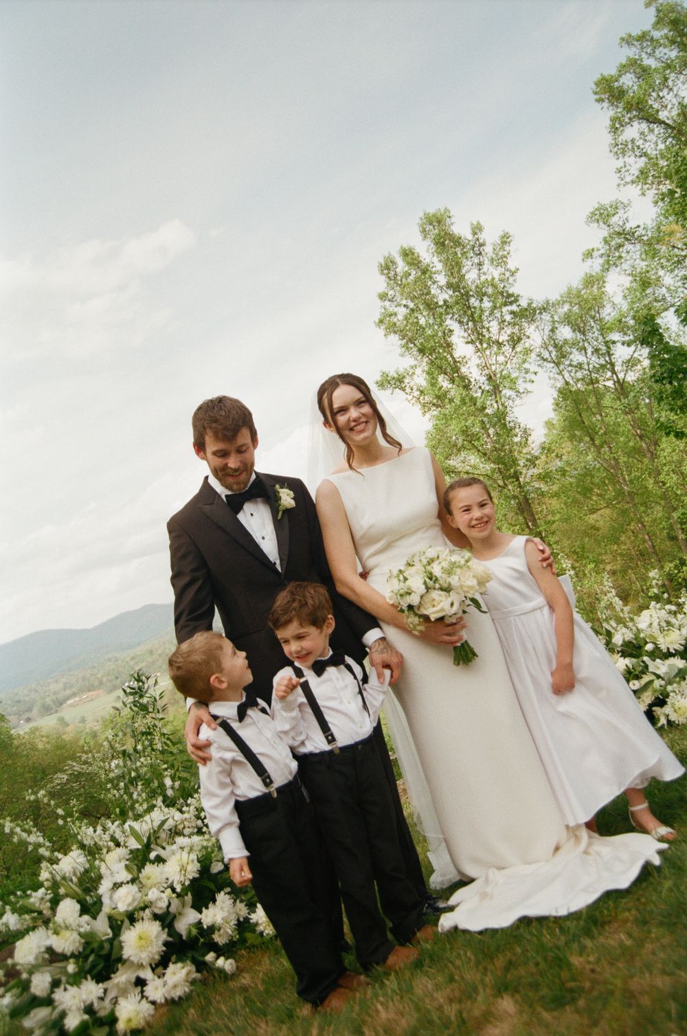 Bride and groom pose with two children in wedding attire on a green hillside with mountain views and white flowers.