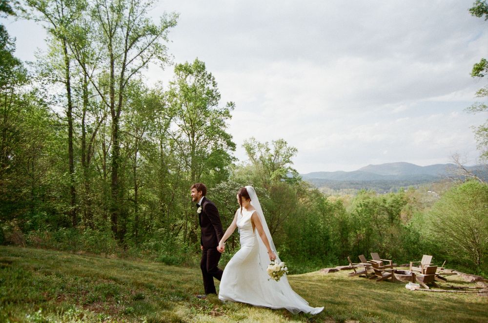 Bride and groom walk hand in hand through lush green hillside with mountain views on wedding day.