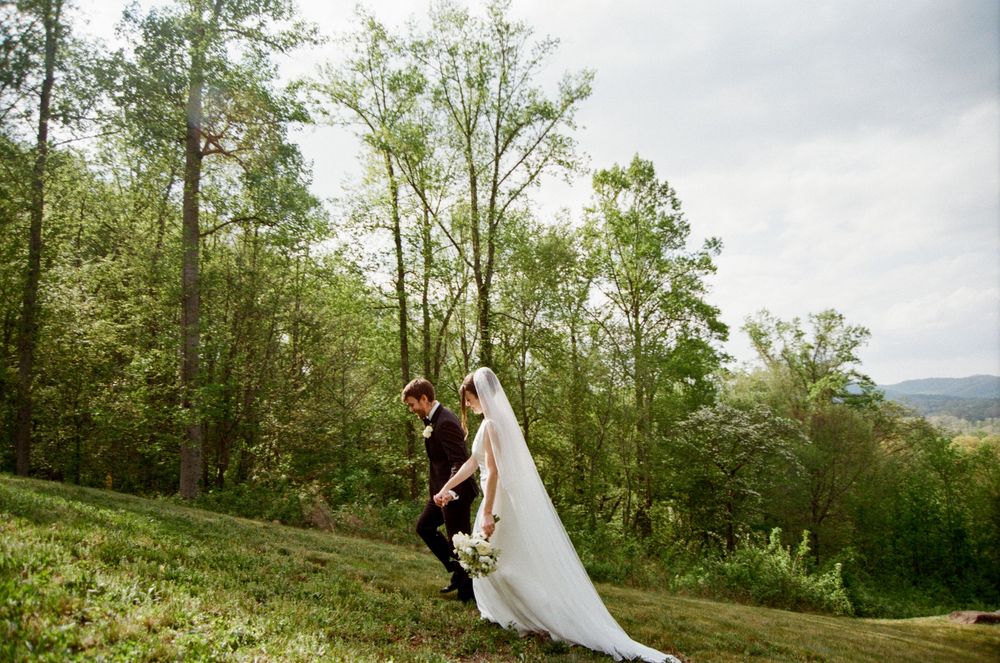 Bride in flowing white gown and groom share intimate moment walking on grassy hill at wedding.