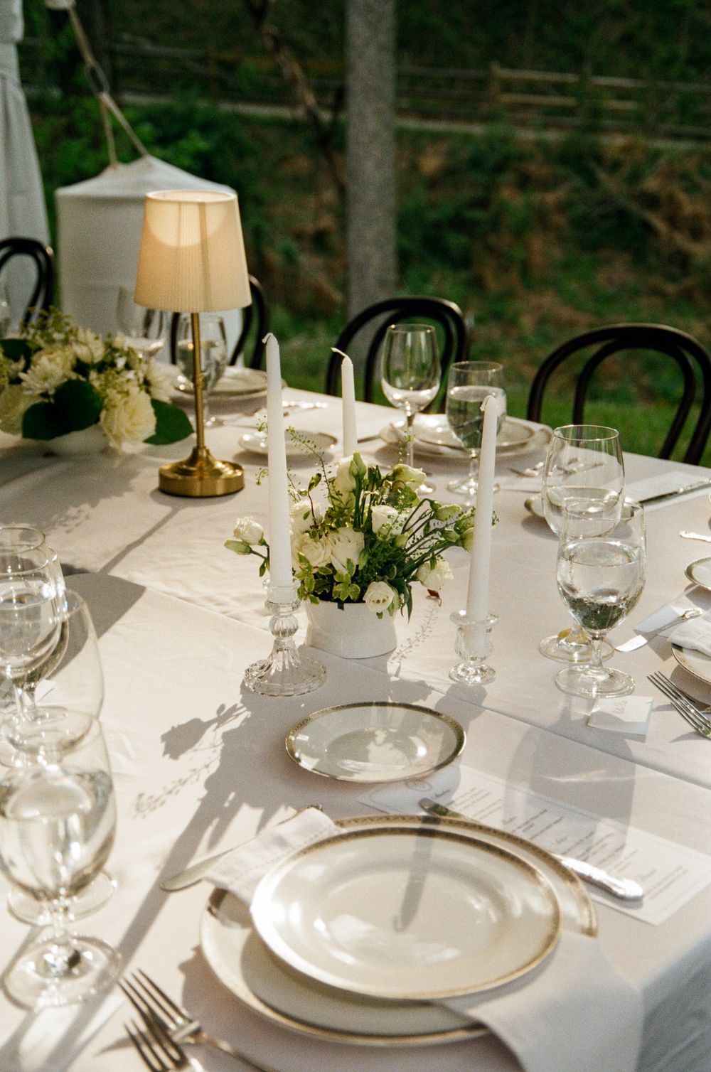 Formal white table setting with gold-rimmed plates, crystal glasses, white candles, and floral centerpiece outdoors.