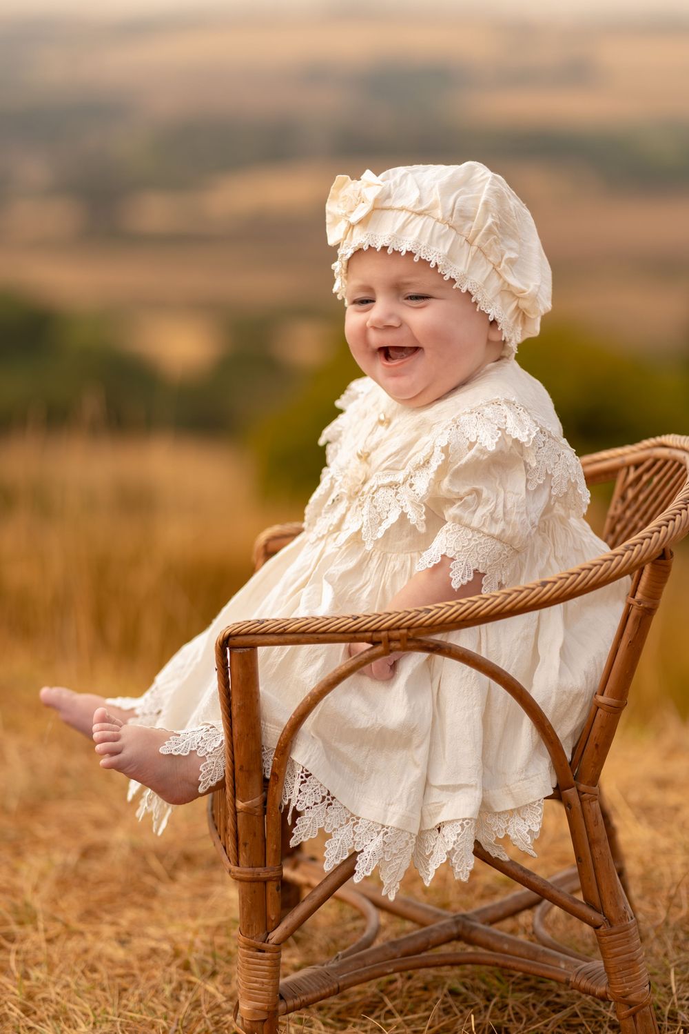 A laughing baby in a cream lace dress and bonnet sits in a wicker chair outdoors in a golden field.