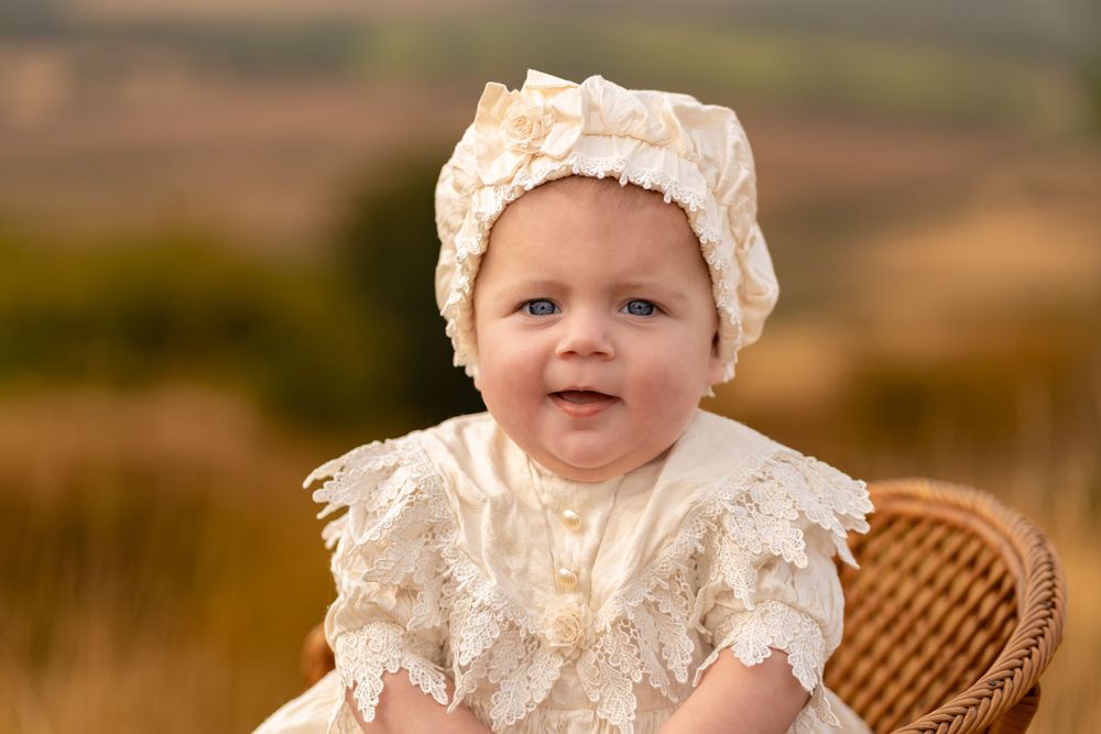 Smiling baby in cream lace dress and bonnet sitting in wicker chair outdoors in golden field.