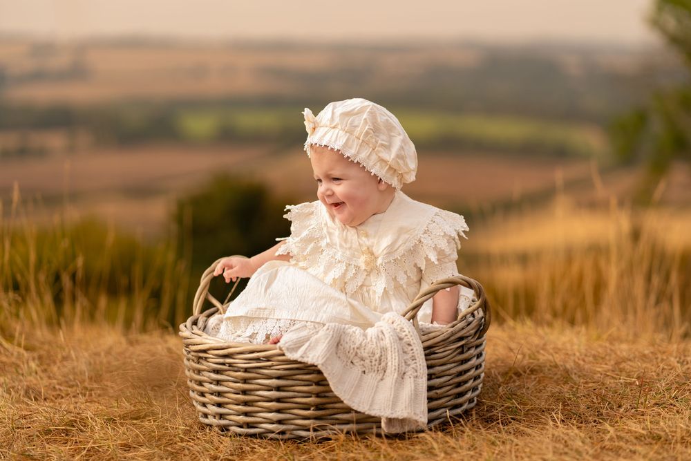Adorable baby in white lace dress and bonnet sitting in wicker basket outdoors in golden field.