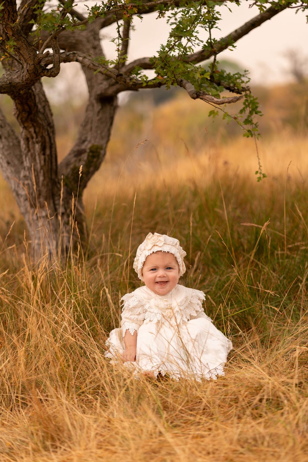 Smiling baby in white dress and bonnet sitting in golden grass field beside a tree.