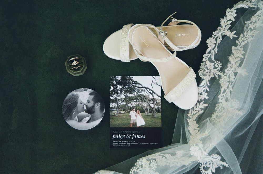 White bridal heels, wedding invitation for Paige and James, lace veil, and a photo on a dark green velvet surface.