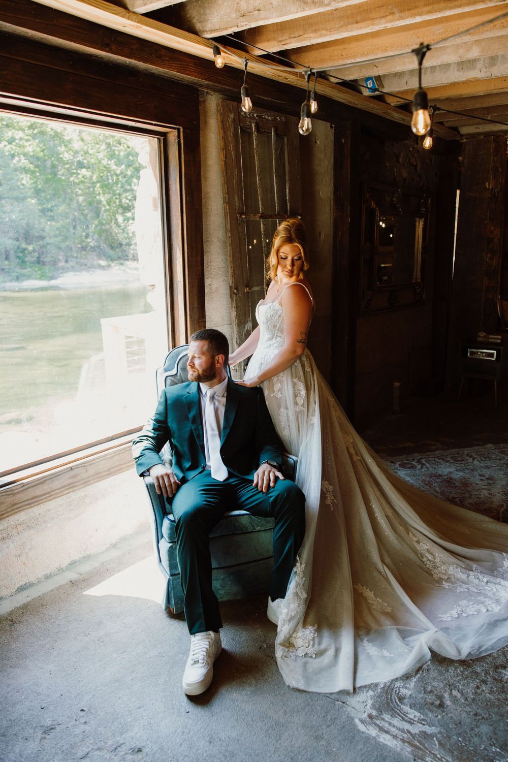 Bride in lace gown stands behind groom in teal suit seated in velvet chair inside rustic barn venue.