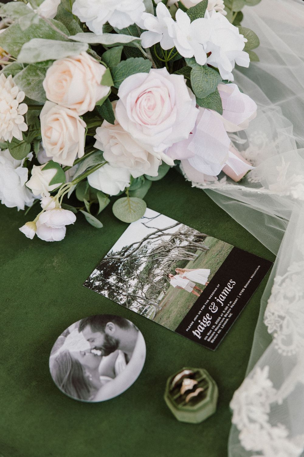 Wedding flat lay with white roses bouquet, bridal veil, save-the-date card, and couple photo button on green velvet.
