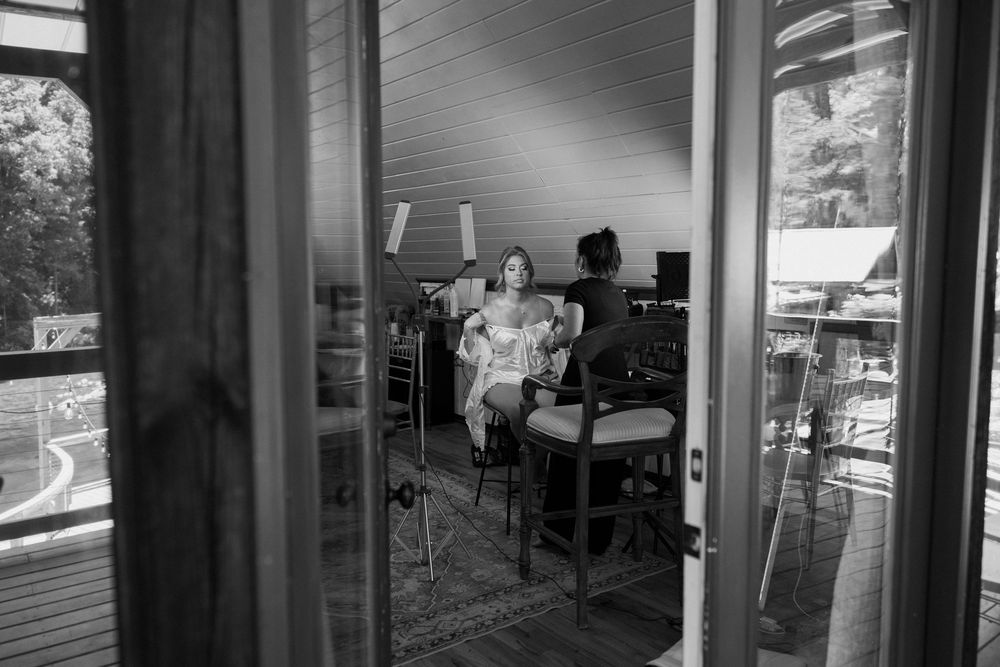 Black and white photo of a bride getting ready in a sunlit room, reflected through glass doors.