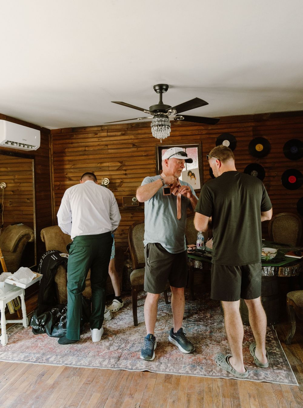 Three men in casual attire gather in a rustic wood-paneled room with a ceiling fan, appearing to prepare for a formal event.