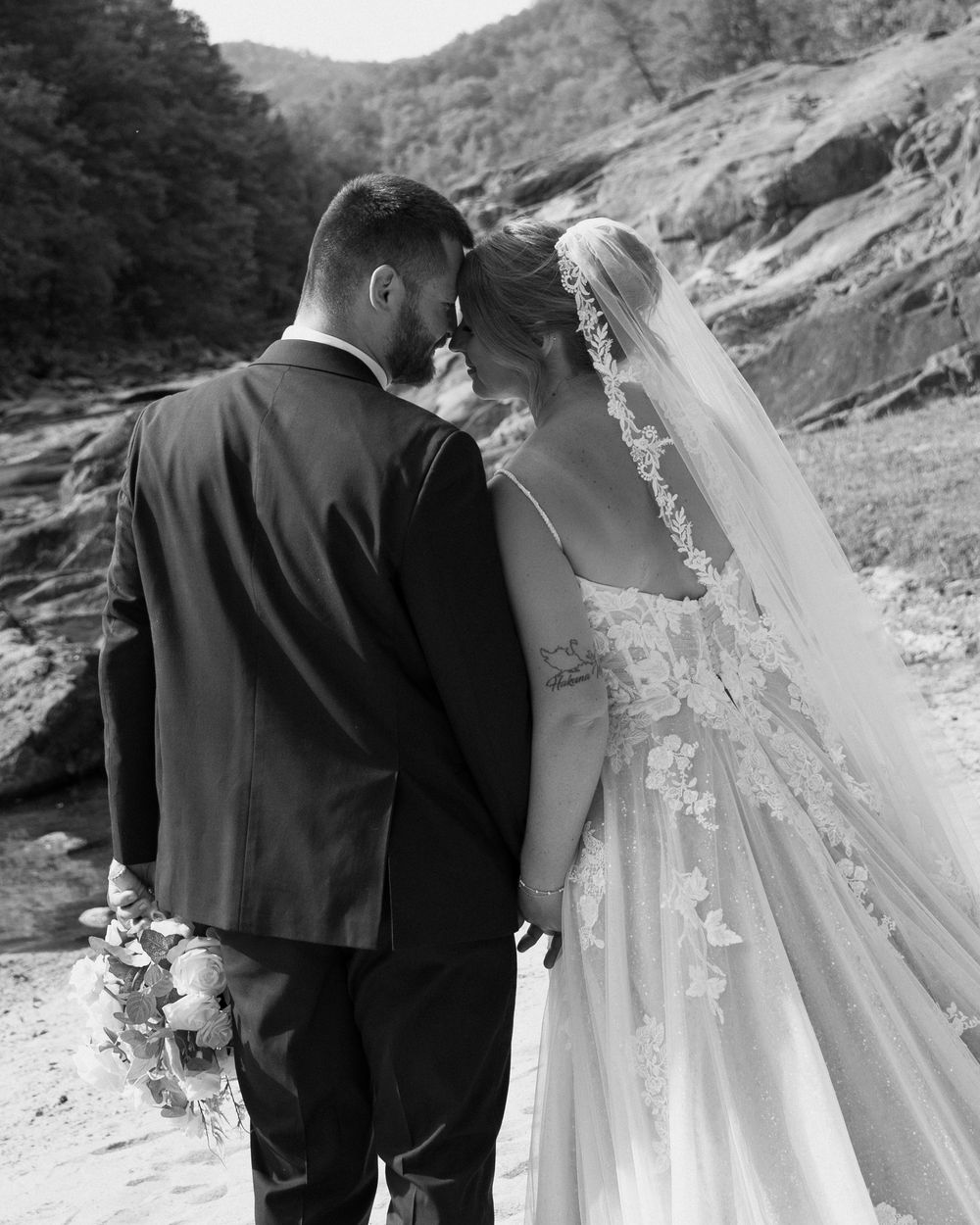Black and white photo of a bride and groom touching foreheads outdoors near rocky terrain on their wedding day.
