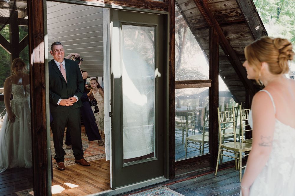 Groom smiles as bride approaches through glass doors at rustic outdoor wedding venue.