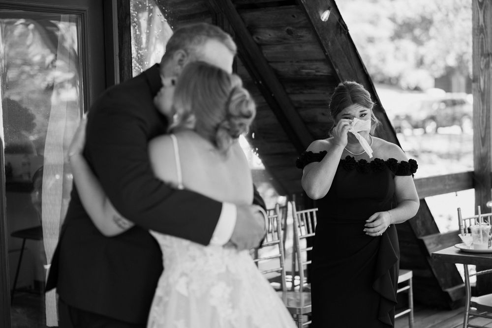 Black and white wedding photo of couple embracing while emotional woman in black dress wipes tears nearby.