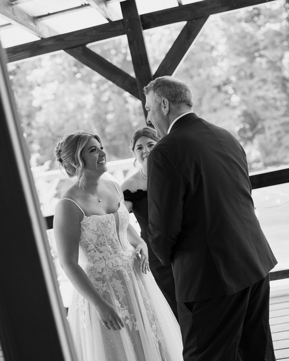 Black and white wedding ceremony photo of smiling bride in lace gown exchanging vows with groom under rustic pavilion.