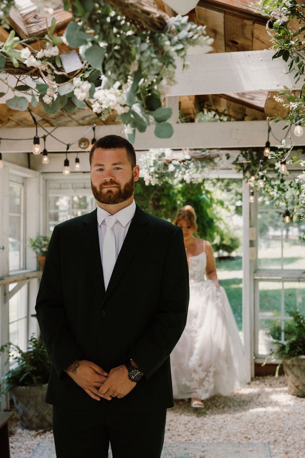 Groom in black suit stands inside floral-decorated wedding venue as bride in white gown approaches from behind.