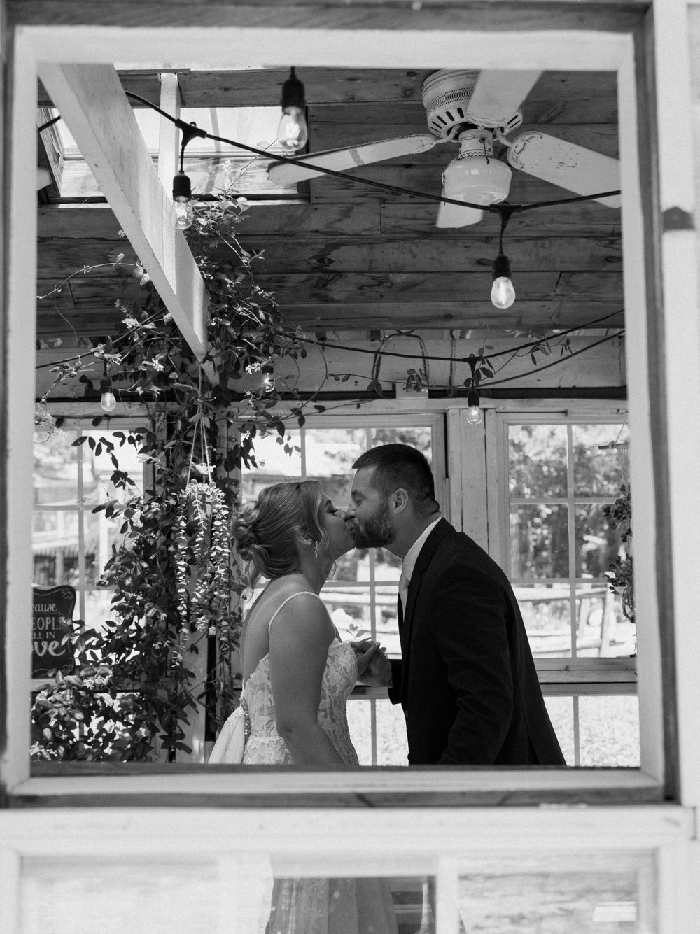 Black and white photo of a bride and groom sharing a kiss inside a rustic venue framed by a window.