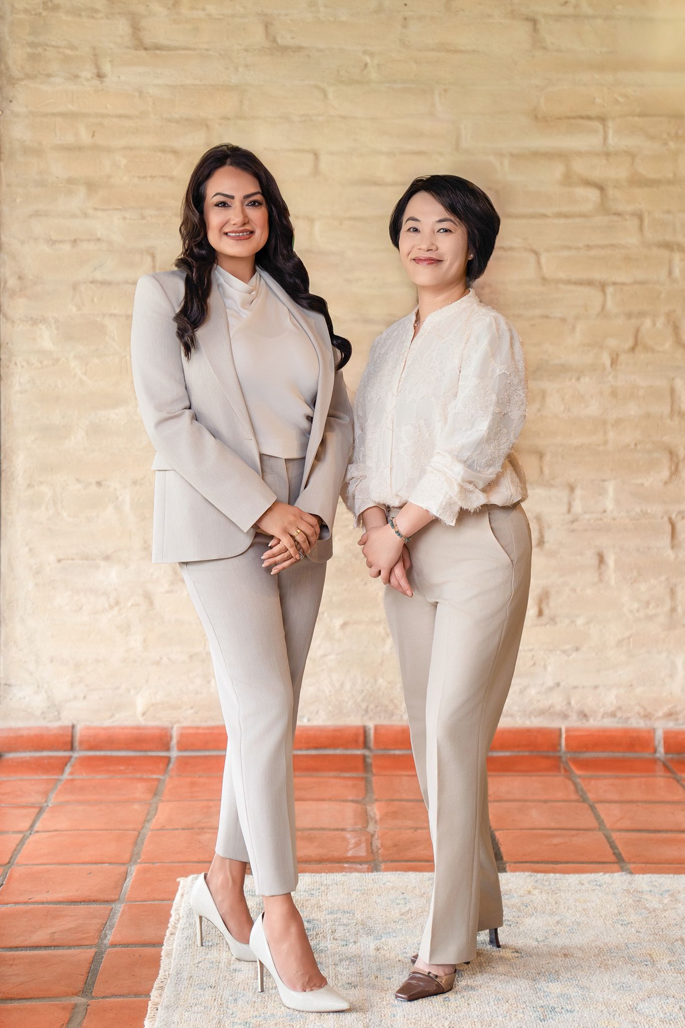 Two professional women in neutral business attire posing together against a brick wall on a tiled floor.