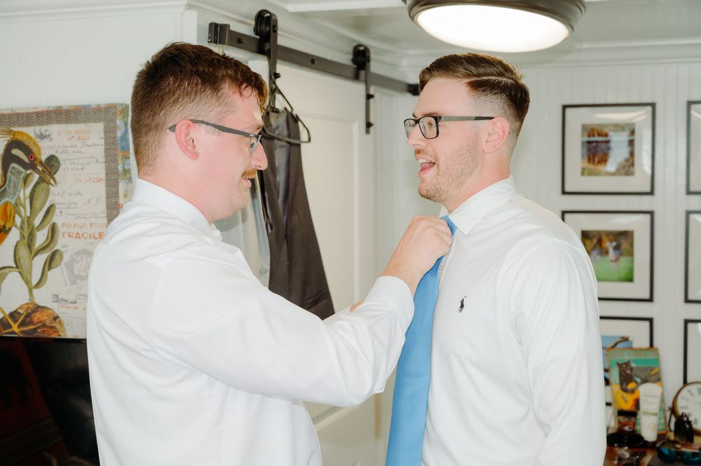 Two men in white shirts, one adjusting the other's blue tie before a wedding.